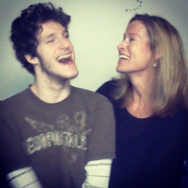 A young man and an older woman sharing a joyful moment, laughing and smiling at each other against a plain background.