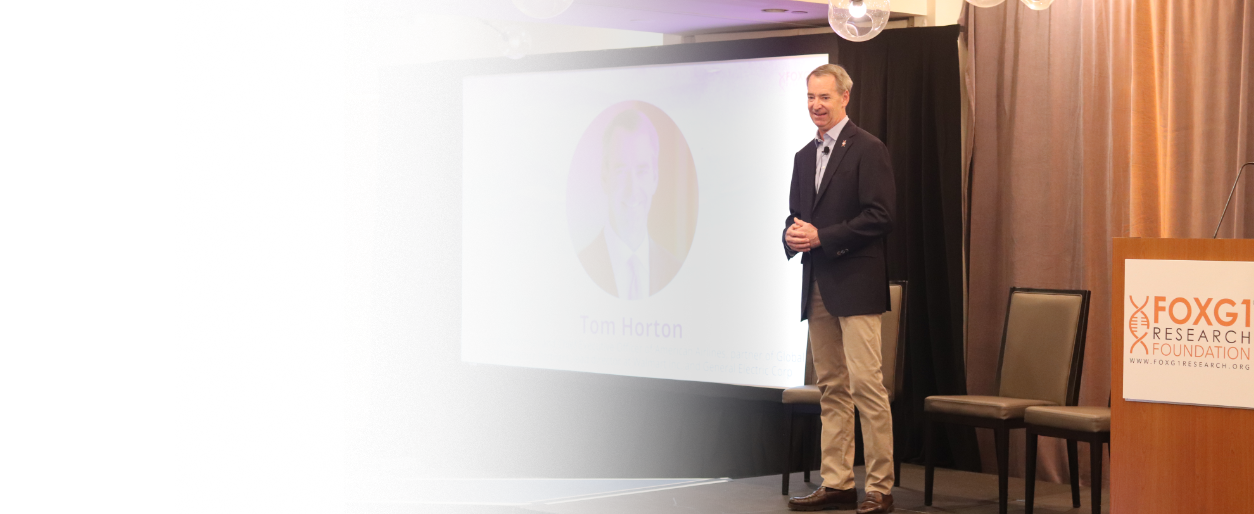 A man in business attire standing on stage at a conference, with a presentation slide displaying his photo and name 'Tom Horton'. The stage has a podium with a sign reading 'FOXG1 Research Foundation' and some chairs against a curtain backdrop.
