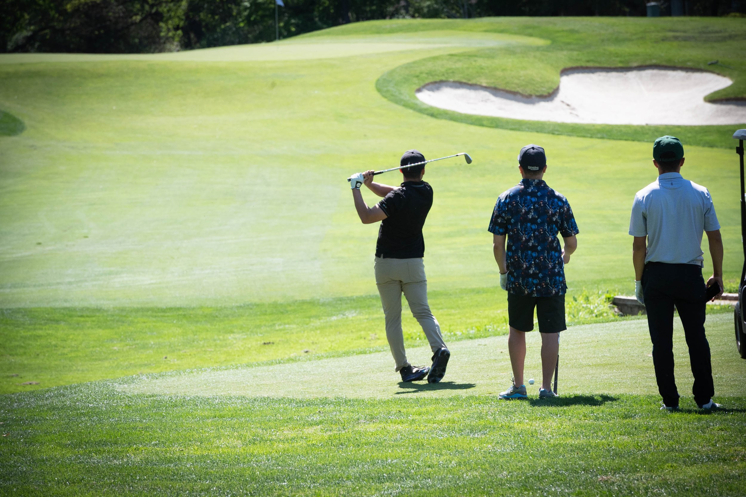 Three people on a golf course with one person about to hit a golf ball towards the green, while the other two watch.
