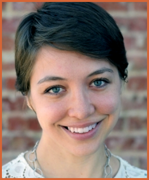 A young woman with short brown hair and blue eyes smiling outdoors against a brick wall.