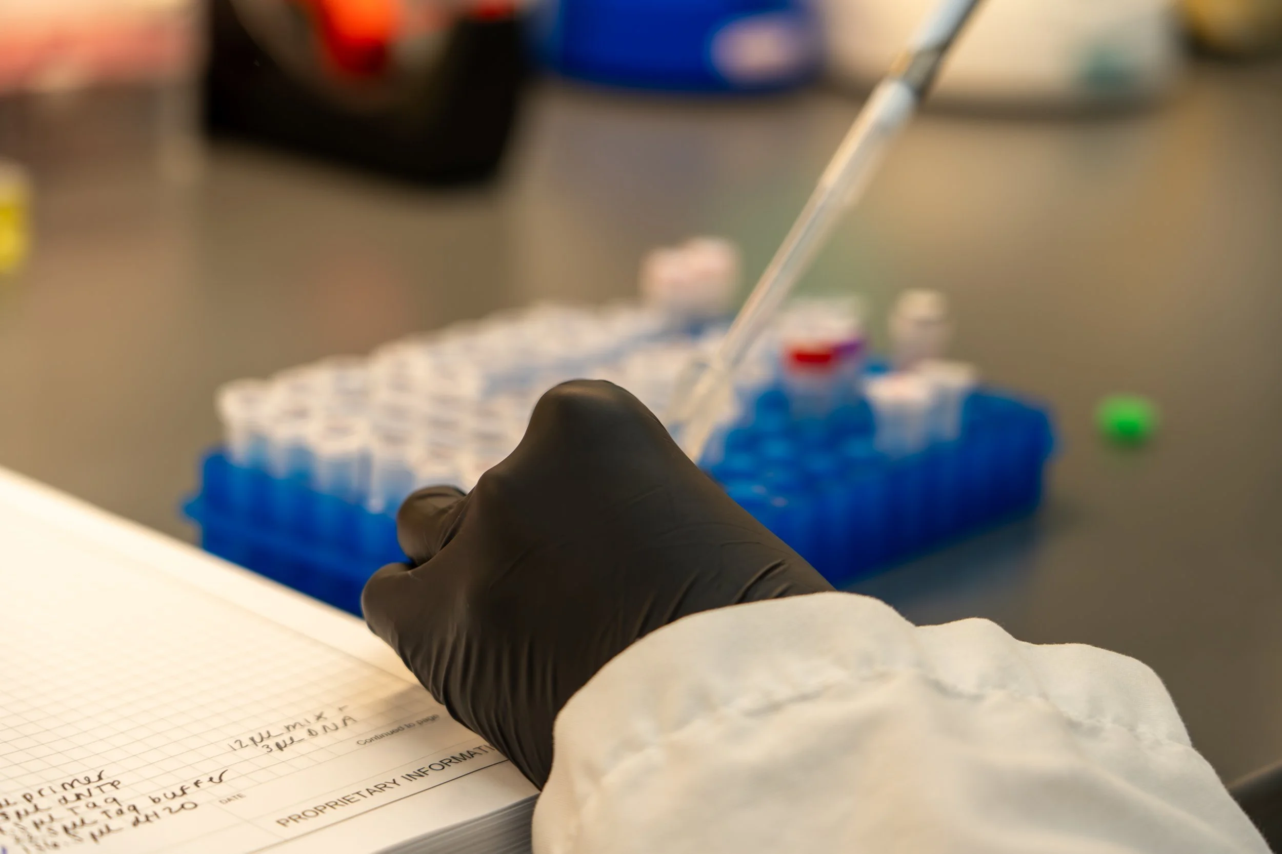 A scientist or lab technician wearing a black glove and white lab coat pipetting liquid into a test tube in a laboratory.