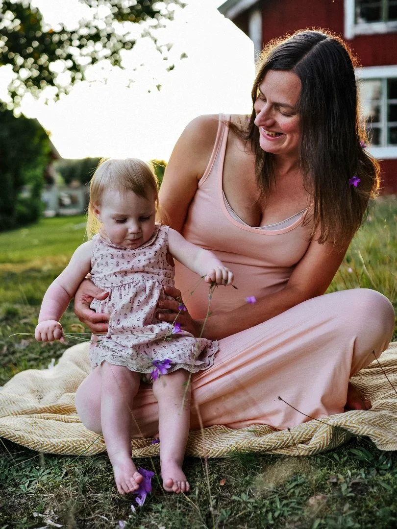 A woman and a young girl with FOXG1 syndrome sitting on a blanket outdoors, playing with purple flowers at sunset in a grassy yard with trees and a house in the background.