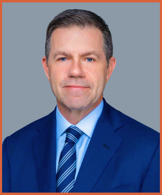 A professional headshot of a middle-aged man with short dark hair, blue eyes, wearing a navy suit, light blue shirt, and a striped blue tie, against a gray background.
