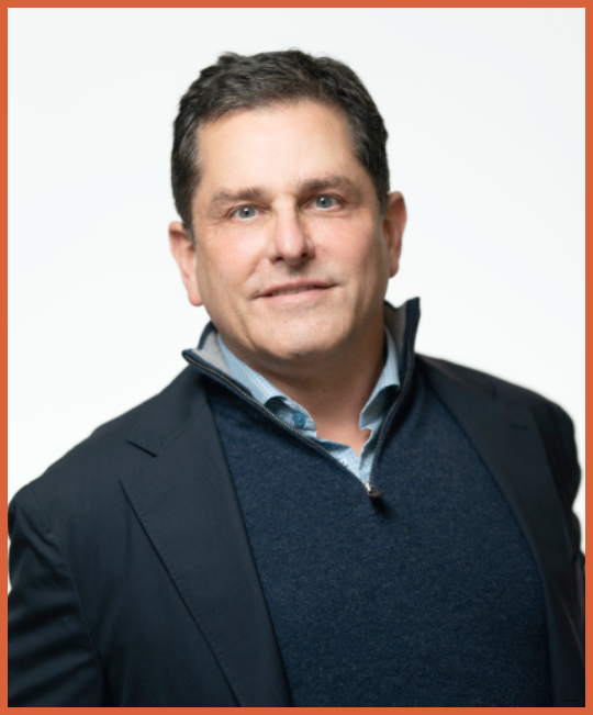 Headshot of a man in a dark suit jacket and a blue sweater, smiling slightly, with a light background and a burnt orange border at the top.
