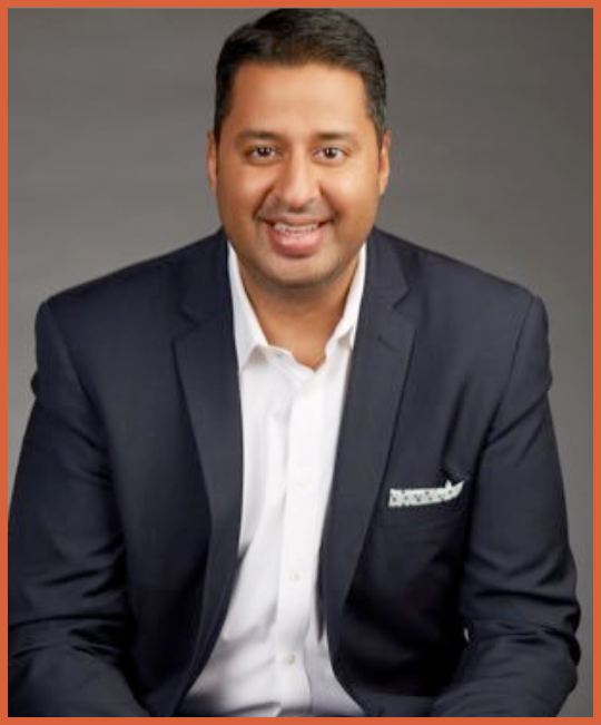 Portrait of a man in a navy blazer and white shirt, smiling in front of a plain gray background.