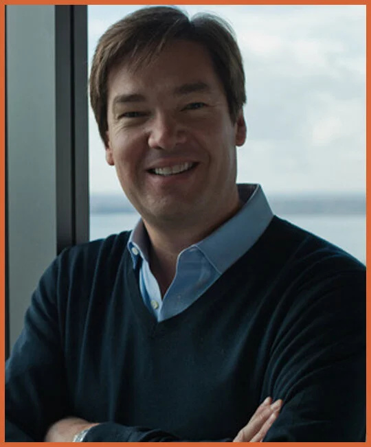 A smiling man with light brown hair wearing a blue collared shirt and navy sweater, standing by a window with water and sky in the background.