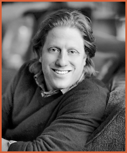 Black and white photo of a smiling man with shoulder-length hair, sitting on a couch.