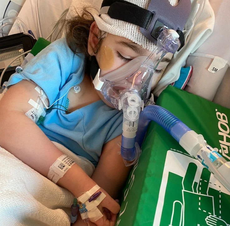 A young girl in a hospital bed asleep, wearing a headband with a breathing mask attached, IV lines, and hospital wristbands, with medical equipment around her.