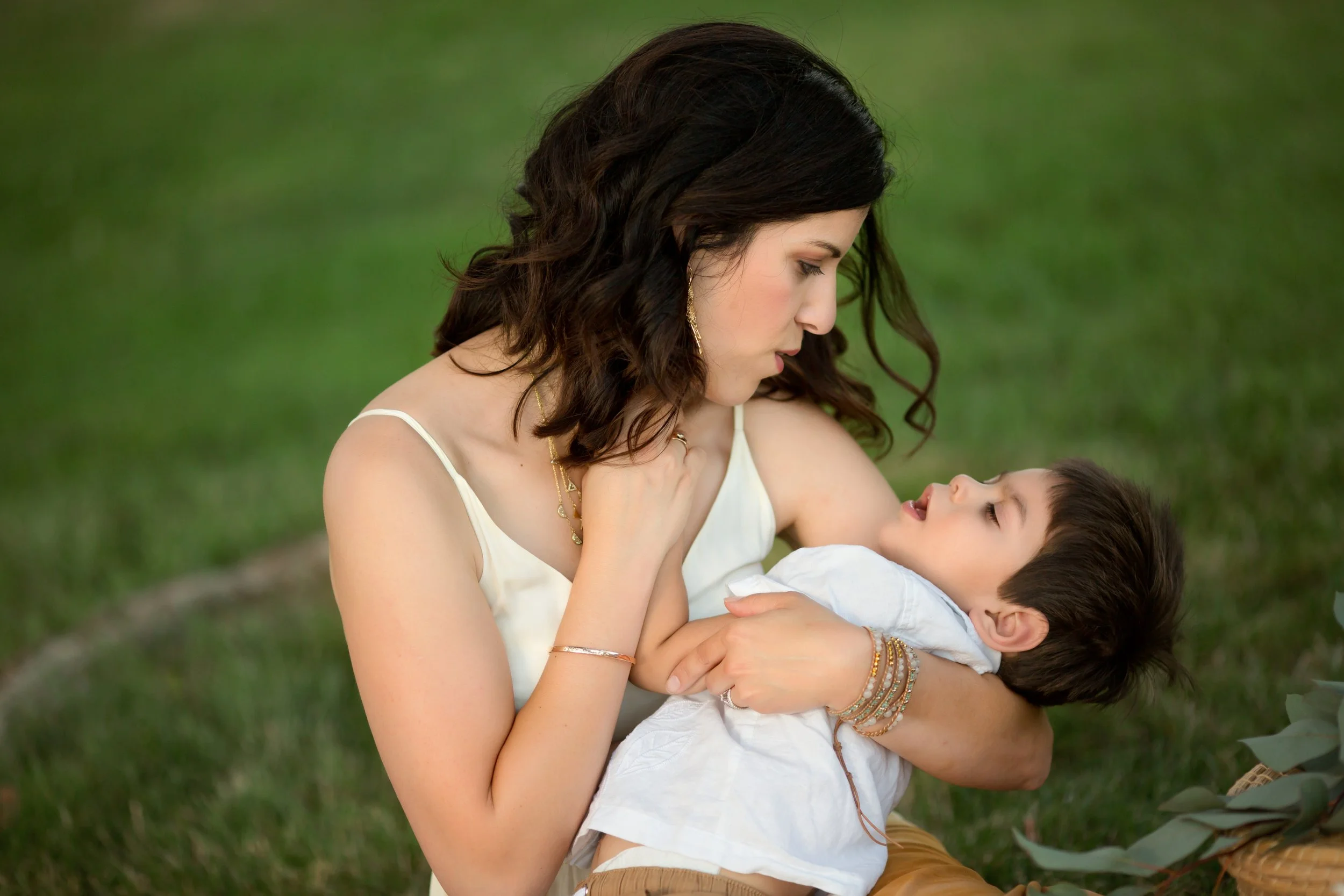 A woman holding a sleeping boy outdoors on a grassy area, looking at him affectionately.