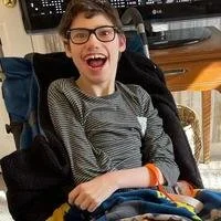 A young boy with FOXG1 syndrome wearing glasses sitting in a wheelchair, smiling, in an indoor setting.