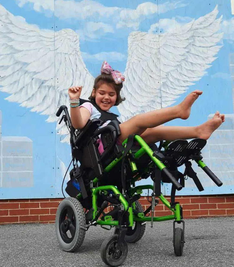 Young girl with a pink bow in her hair sitting in a green and black wheelchair, smiling with her legs raised, in front of a mural of white angel wings on a light blue background.