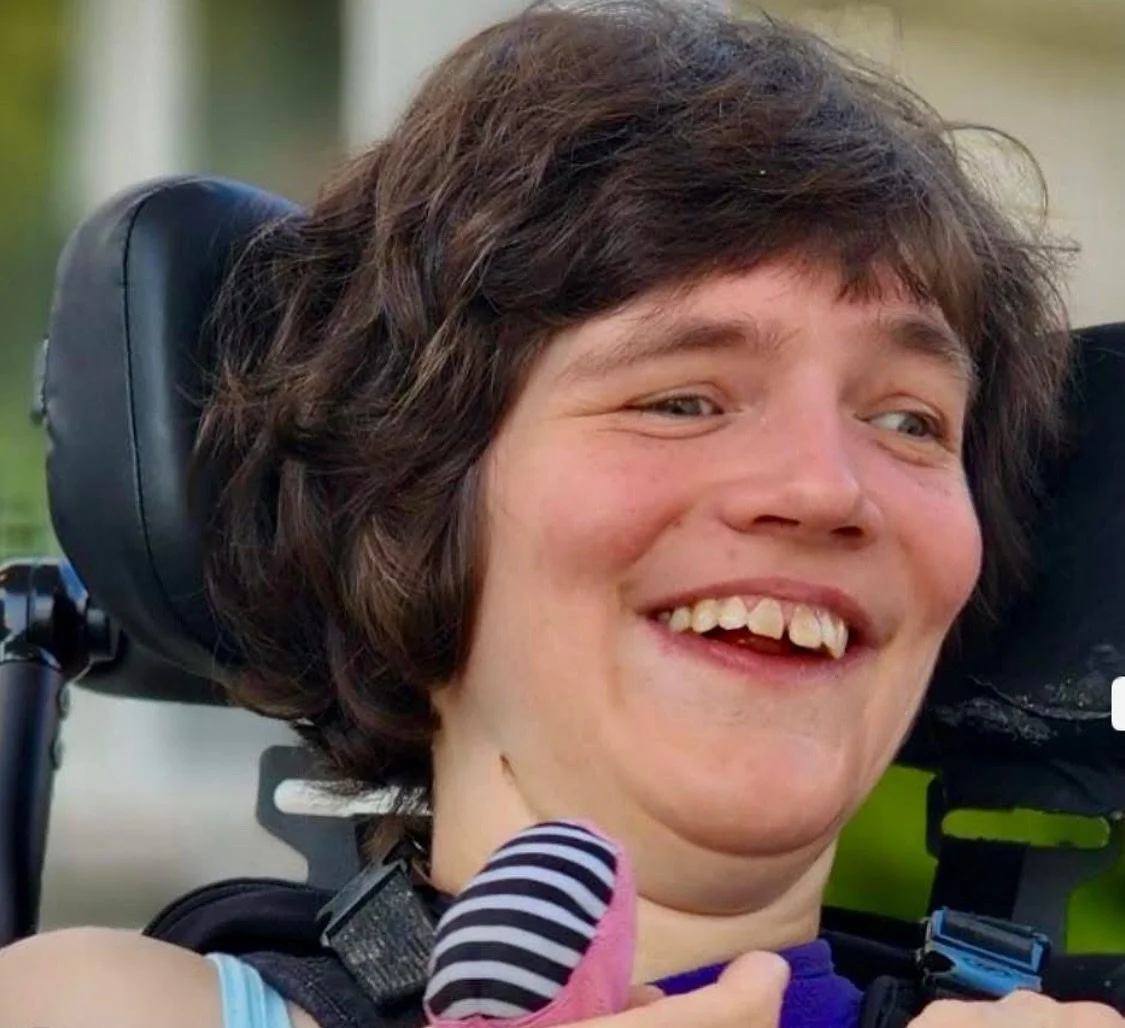 A young woman with FOXG1 syndrome with short brown hair smiling, sitting in a wheelchair.