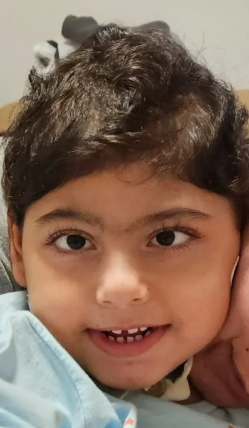 A young boy with dark, curly hair and big brown eyes smiling showing missing front teeth, lying close to an adult, possibly a parent, with part of their face visible, in an indoor setting.