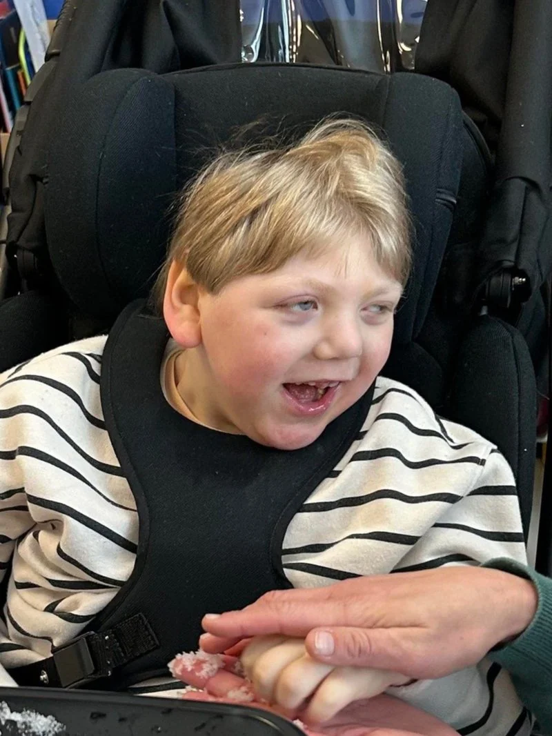A young boy with blond hair in a striped shirt sitting in a wheelchair, smiling and holding hands with an adult who is offering him a piece of bread or pastry.