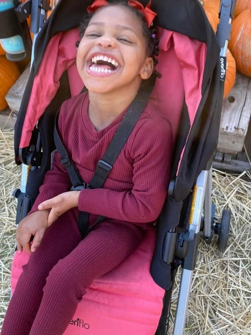 A smiling young girl with curly hair and a red bow, sitting in a pink and black stroller, outdoors among pumpkins and hay.