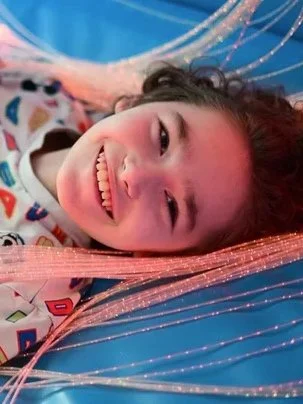 A young girl lying down and smiling, with long pink hair, on a blue background with some decorative strings or beads.