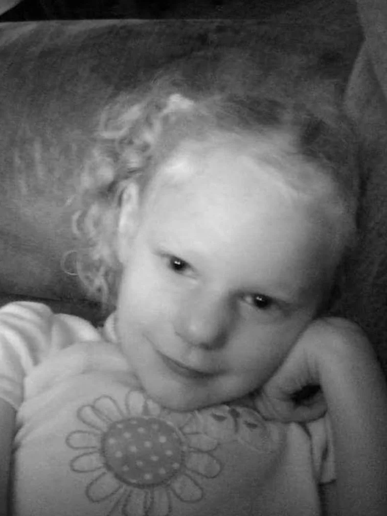 A young girl with curly hair lying on a couch, smiling at the camera, wearing a shirt with a flower pattern.