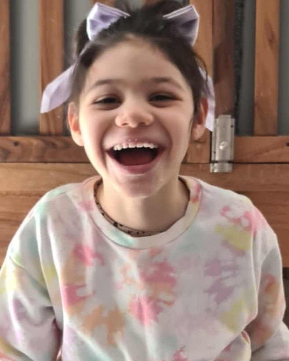 A young girl with short dark hair, smiling widely, wearing a tie-dye shirt and a bow headband, standing in front of a wooden background.