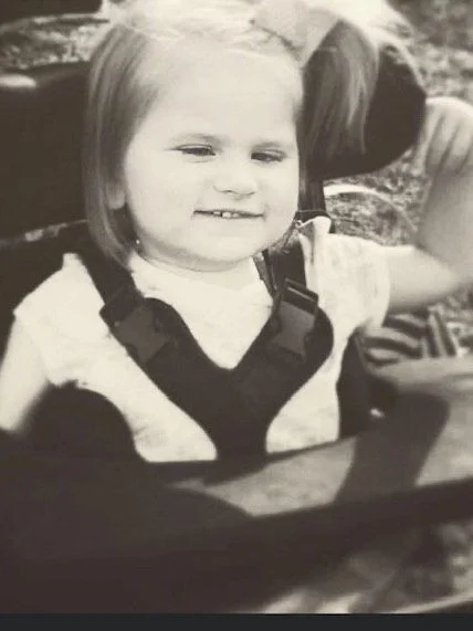 A young girl with short hair smiling, sitting in a stroller outdoors.