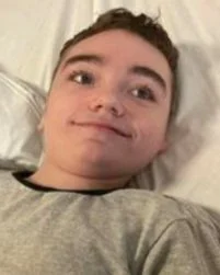 A young person lying on a bed, looking at the camera with a slight smile.