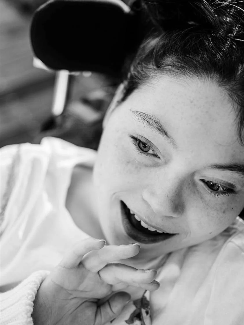 A close-up black and white photo of a smiling young woman with freckles, holding her hand near her face, with a blurred chair in the background.