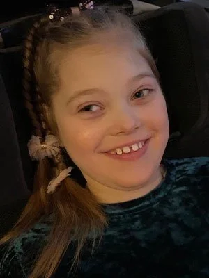 A young girl smiling, sitting in a car seat, with braided hair and wearing a blue patterned shirt.