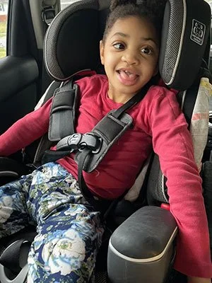 A young girl sitting in a car seat, smiling and sticking out her tongue.