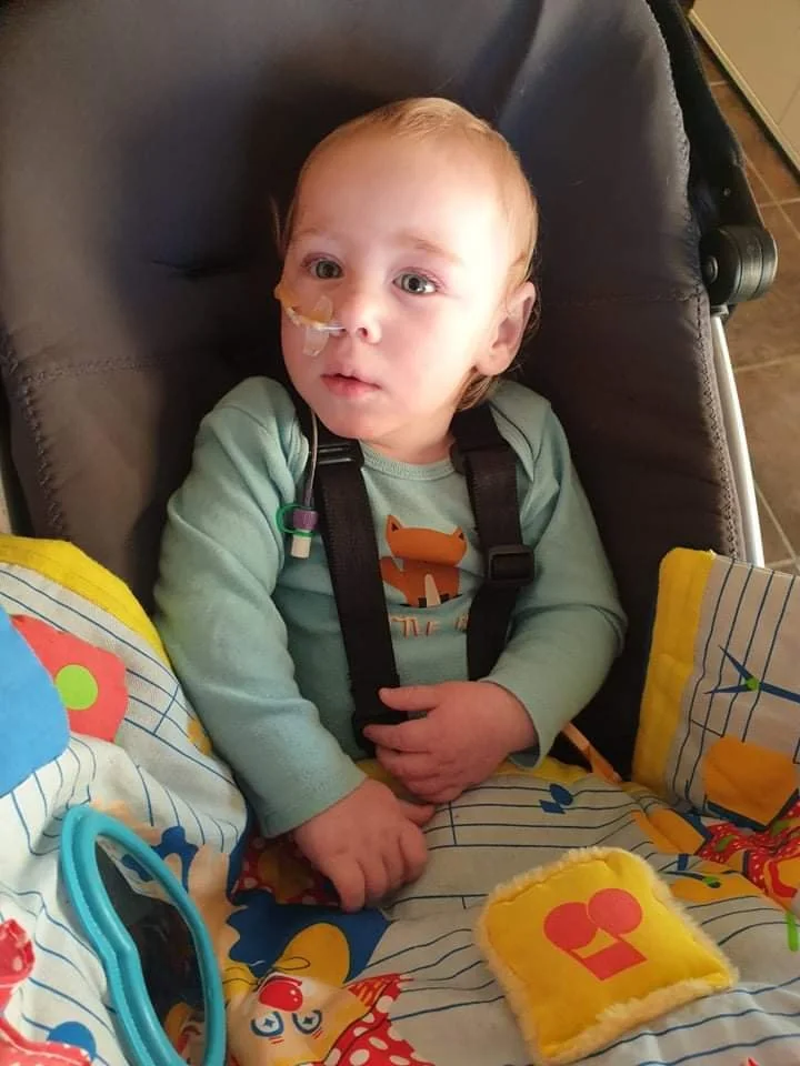 A young child in a wheelchair with a feeding tube in his nose, sitting in a high chair with colorful patterned fabrics, wearing a light green long-sleeved shirt with a fox graphic.