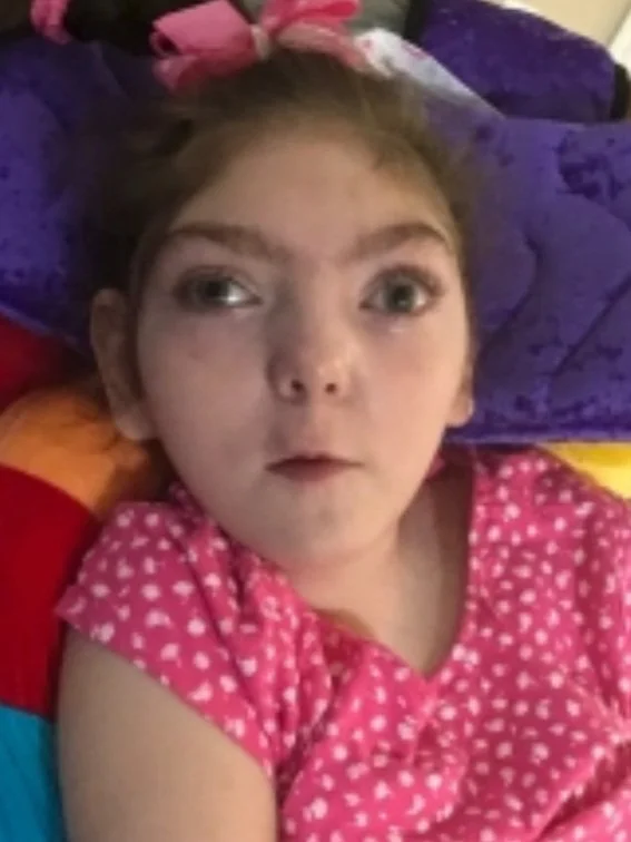A young girl lying on colorful pillows, wearing a pink dress with white polka dots and a pink hairbow.