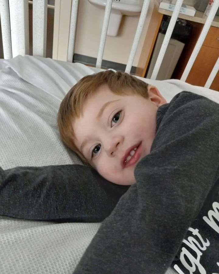Young boy with red hair lying on a hospital bed, resting his head on his arm, smiling softly.