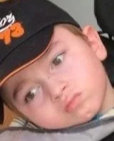 Close-up of a young child's face, wearing a black and orange cap, looking pensively to the side.