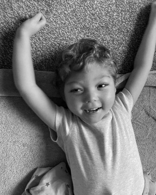 A young child lying on a sofa with arms raised, smiling and looking up, in a black and white photo.