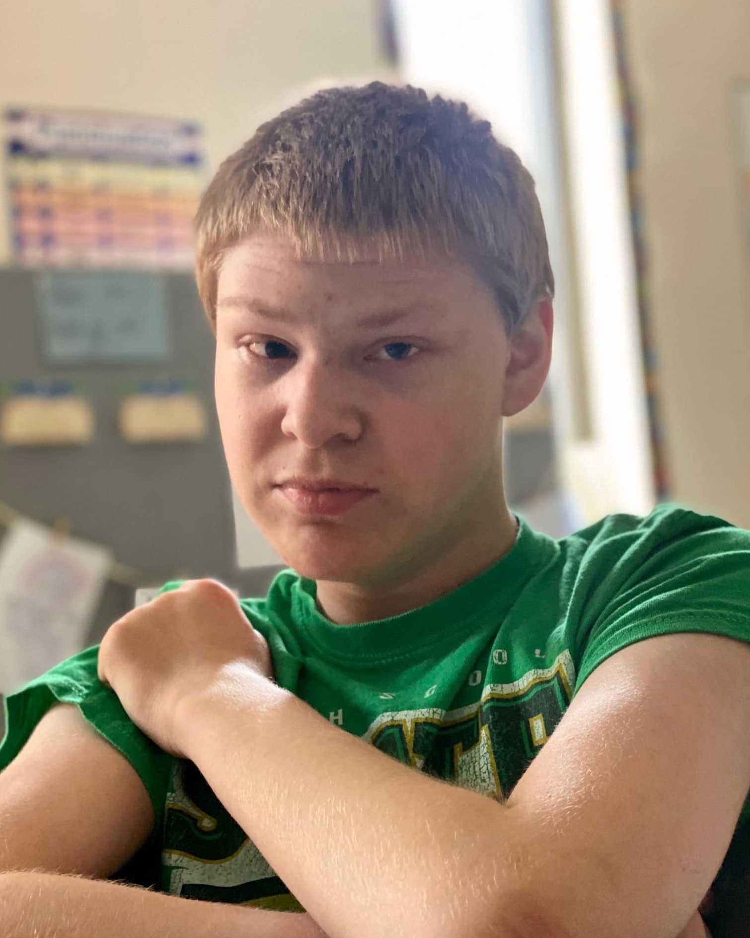 A young boy with short brown hair and fair skin is making a fist, flexing one arm, and looking at the camera. He is wearing a green T-shirt and appears to be inside a room with a blurred background that includes papers or posters on a bulletin board.