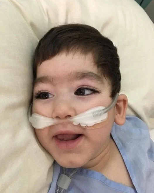 Young boy with a nasal cannula lying in a hospital bed, smiling and looking to the side.