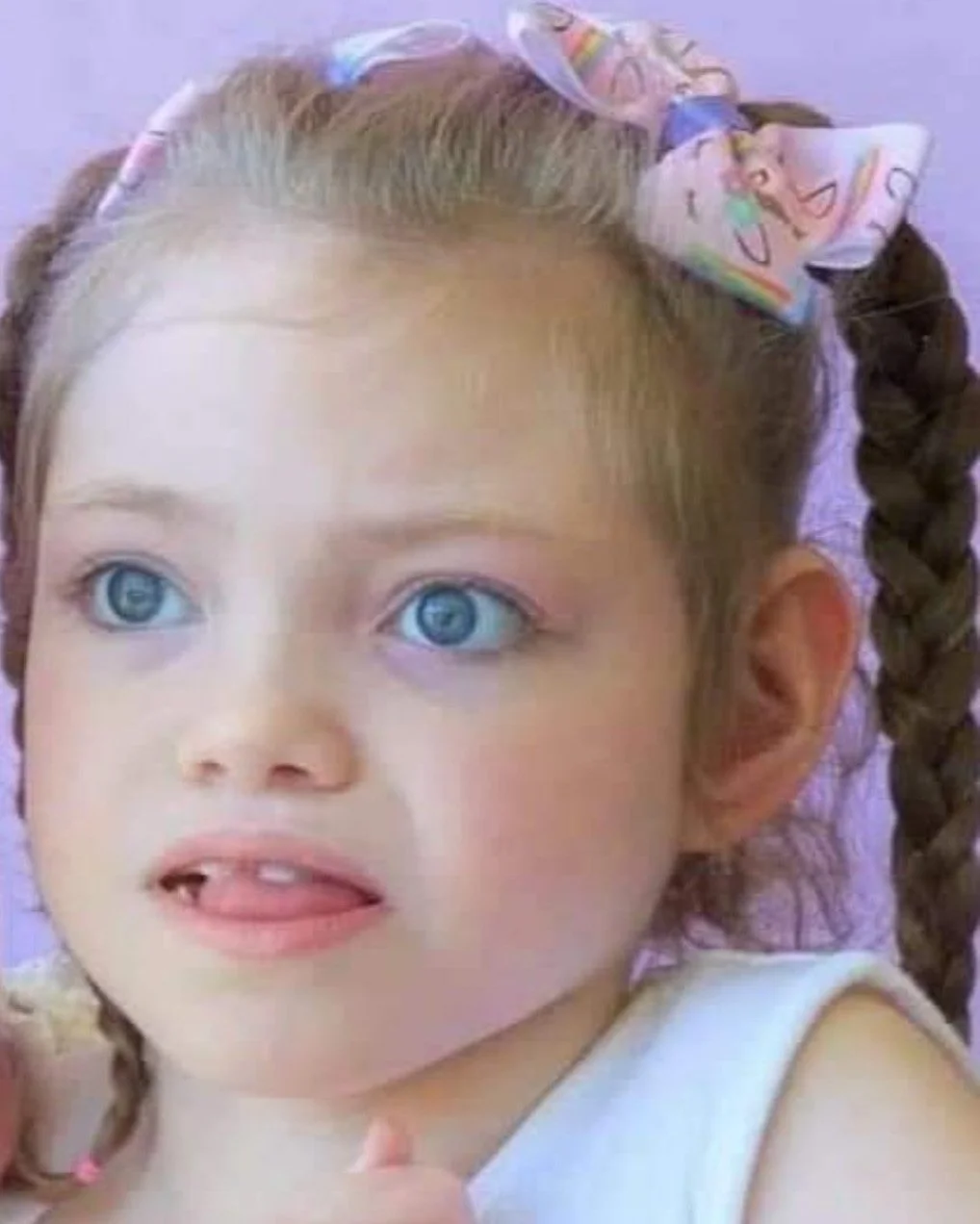 Close-up of a young girl with light blue eyes, light skin, and light brown hair styled into braids with a pastel-colored bow accessory.