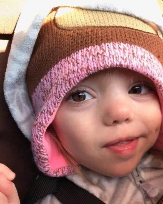 A young child wearing a multi-colored winter hat that is brown, pink, and gray, with a pink lining, smiling with their tongue slightly sticking out.