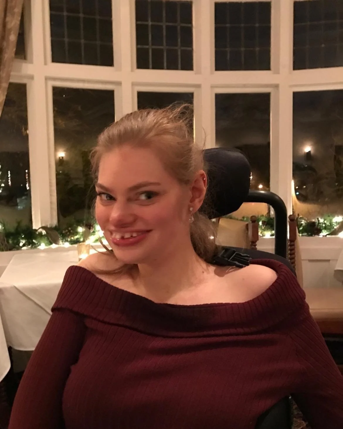 A smiling woman with glasses, wearing a maroon off-the-shoulder top, sitting indoors at a restaurant with large windows and festive decorations.