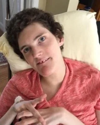A young person with curly brown hair and light skin lying in bed with a pillow behind their head, wearing a red T-shirt.