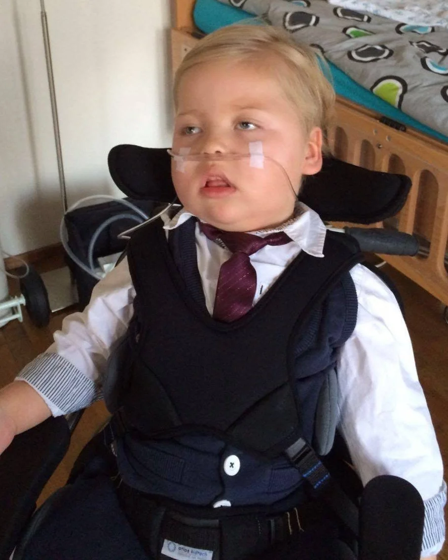 A young boy in a wheelchair wearing a school uniform, including a white shirt, maroon tie, and dark vest, with a nasal cannula for oxygen, in a bedroom with a bed and closet in the background.