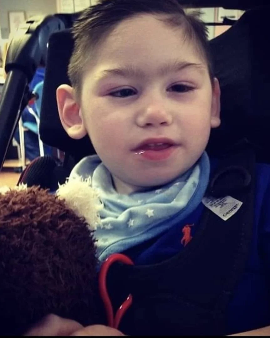 A young boy with short hair sitting in a wheelchair, wearing a blue shirt and bib, holding a brown stuffed animal, with a slight smile and a bit of drool on his lips.