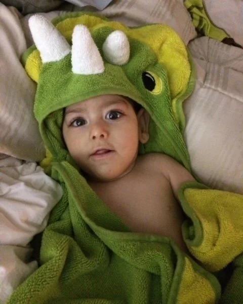 Baby lying on bed, wrapped in a green dinosaur costume with white horns and yellow accents.