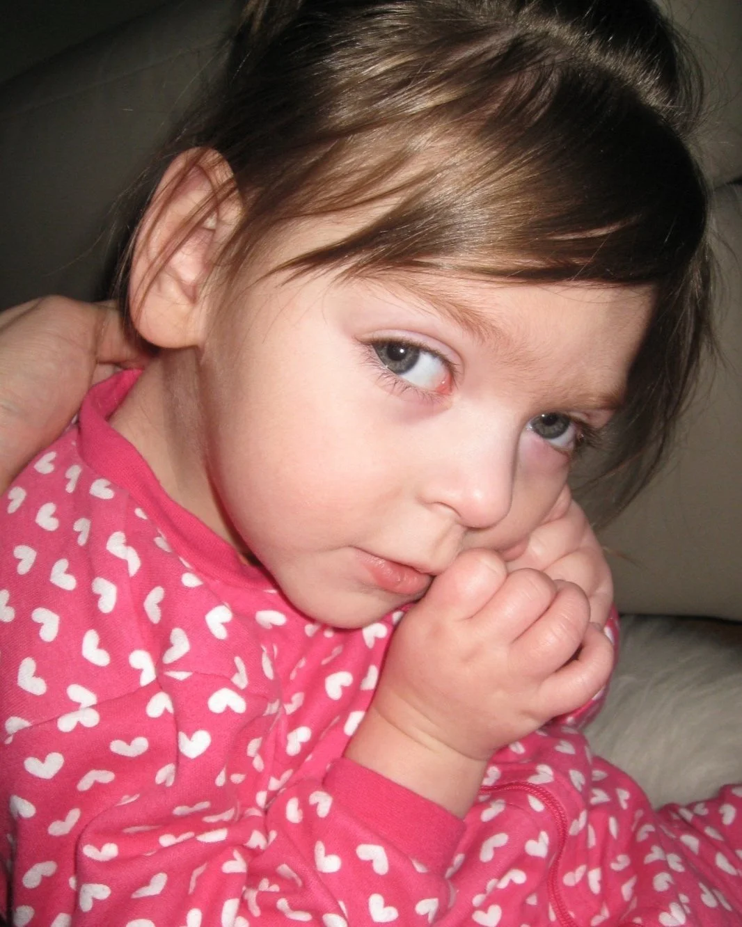 A young girl with brown hair and blue eyes, wearing a pink shirt with white hearts, resting her head on her hand.