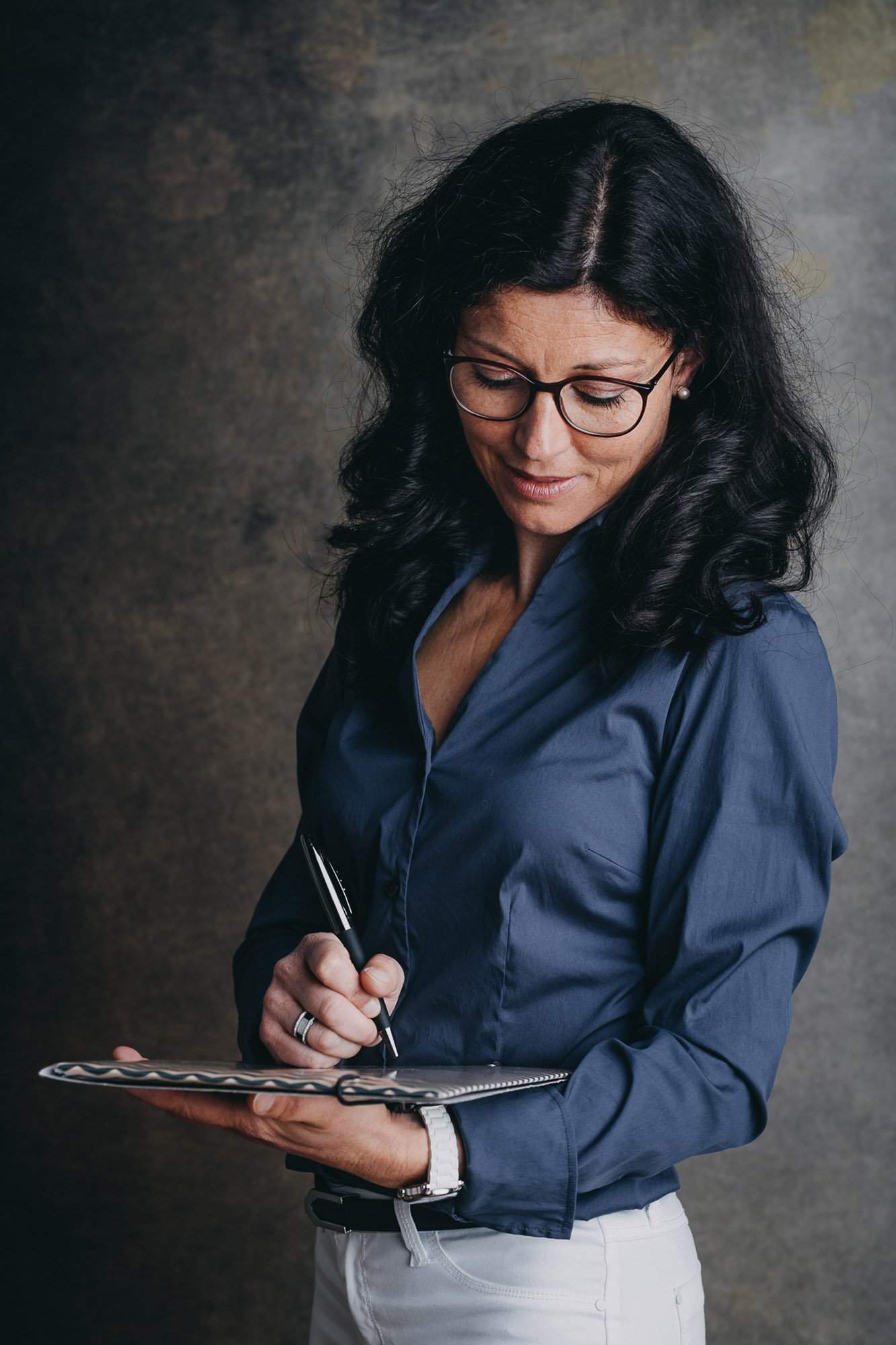 Une femme portant des lunettes noires écrit dans un carnet, vêtue d'une chemise bleue et d'un pantalon blanc, tenant un stylo.