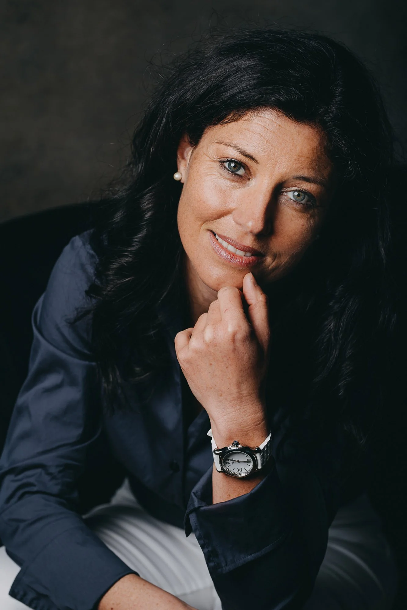 Portrait d'une femme ayant les cheveux noirs, portant une montre noire, un earring en perle, en train de sourire, avec la main sous le menton, habillée d'une chemise bleue, contre un fond sombre.