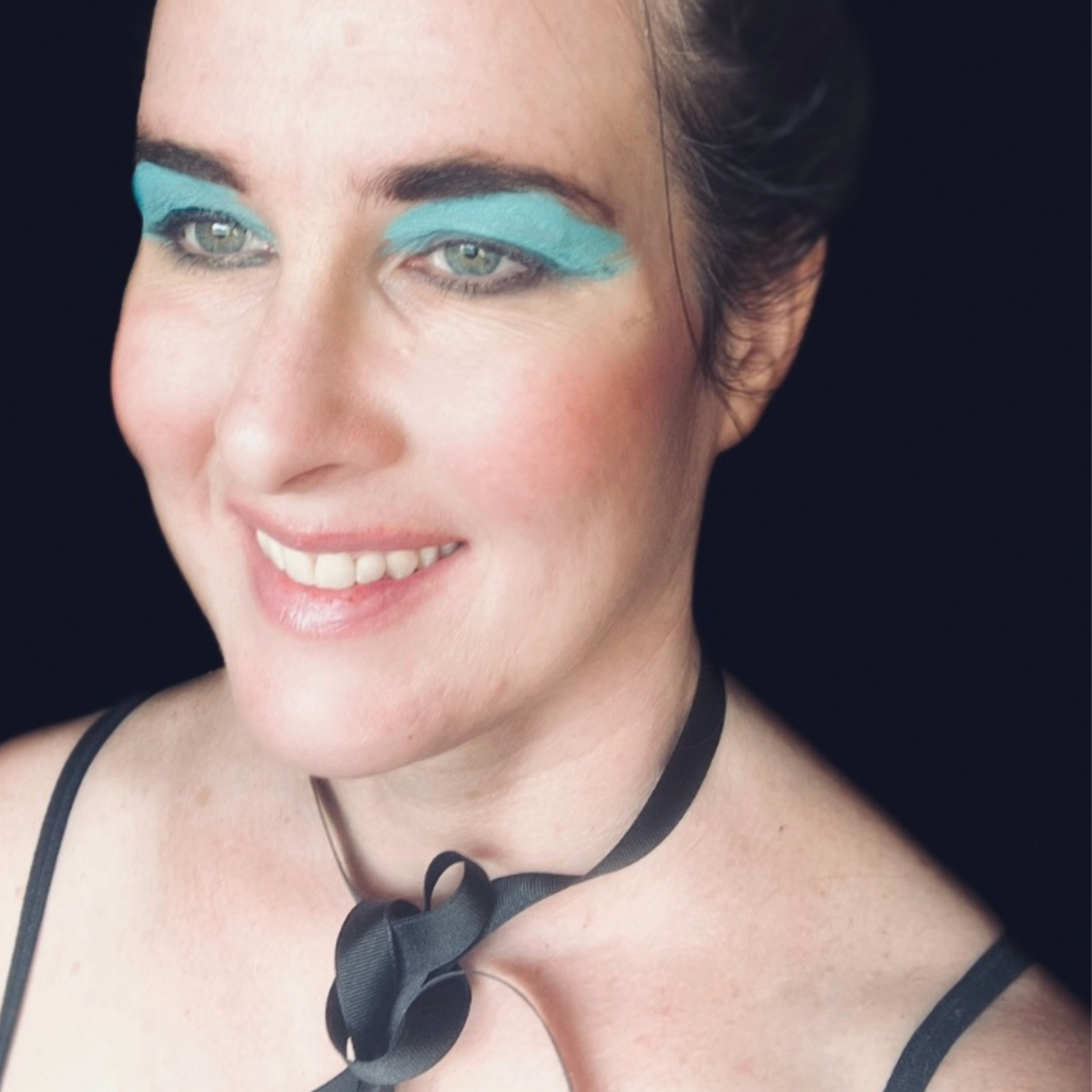 A woman with short brown hair, blue eye makeup, light skin, smiling, wearing a black choker with a bow and black straps, against a black background.