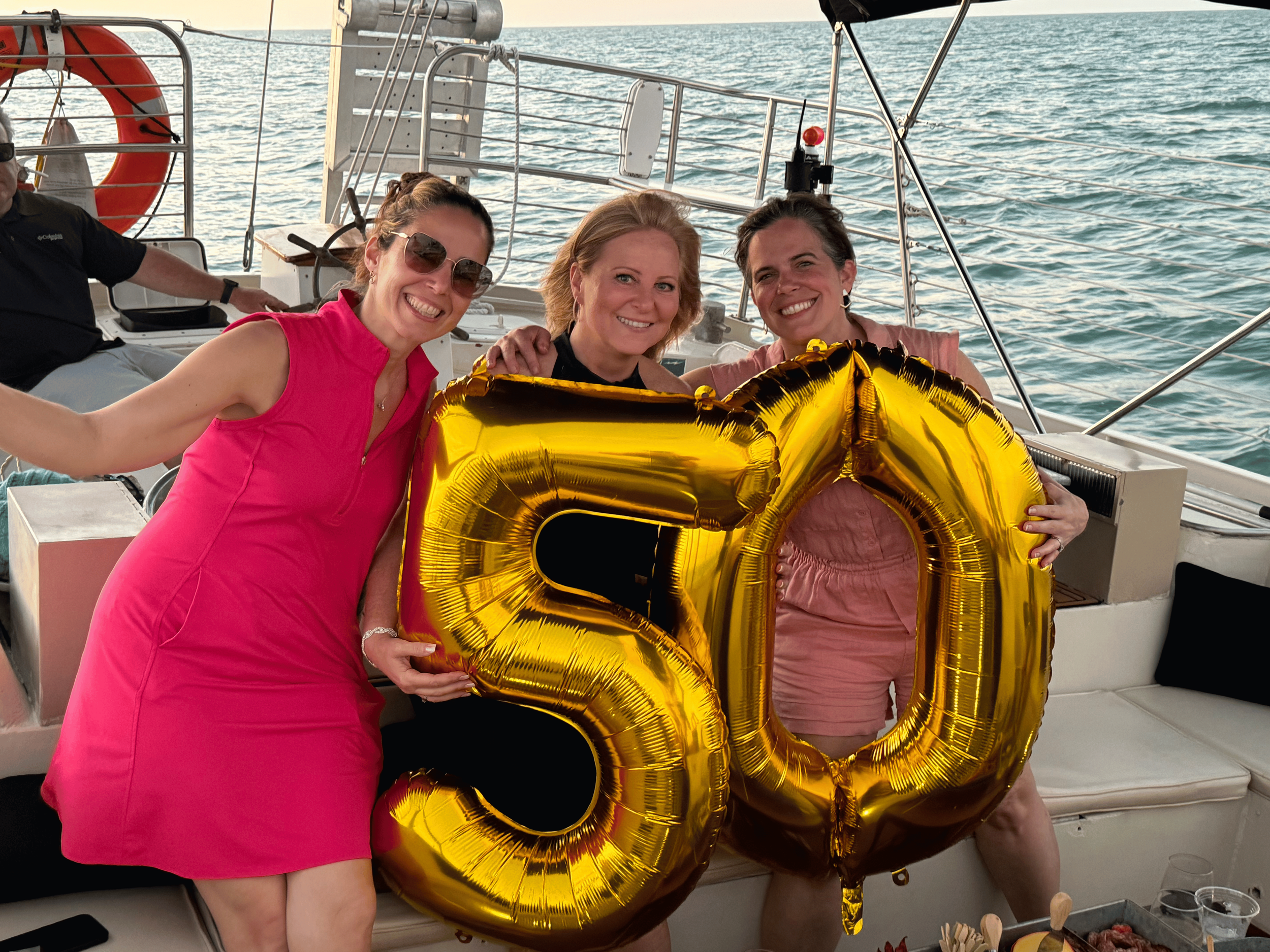 Three women on a boat holding large gold-colored balloons forming the number 50, smiling at the camera with the ocean in the background.