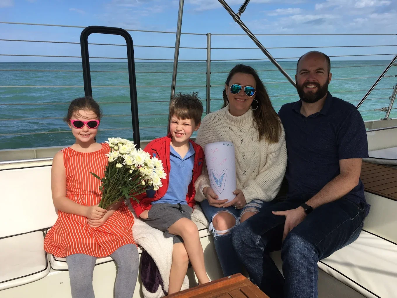 A family of four sitting on a boat with ocean background, holding flowers, an urn, and a handmade card.