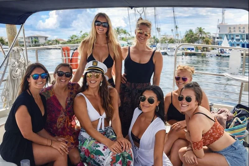 Group of eight women enjoying a boat ride in a marina, wearing summer clothes and sunglasses, with boats and trees in the background.