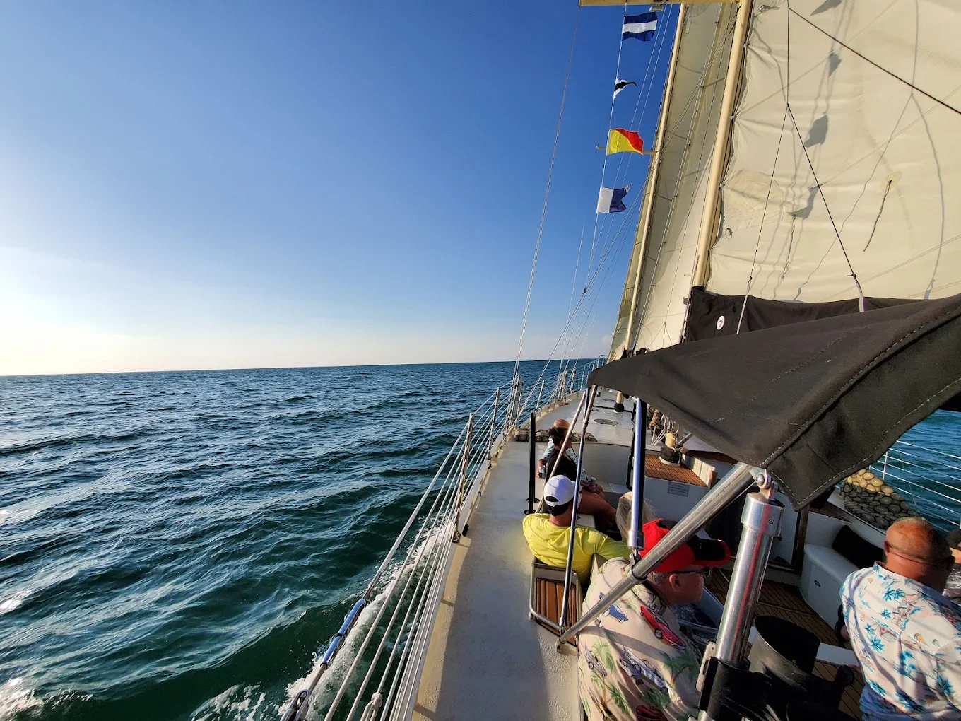 People relaxing on a sailboat with a high mast, sailing on the open ocean under a clear sky, with flags flying from the mast.