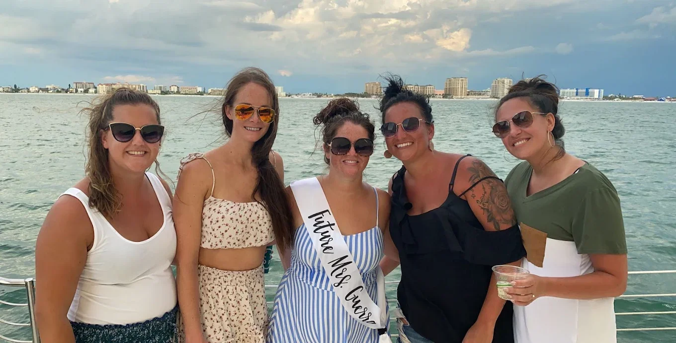 Five women on a boat by the water, one wearing a sash that reads 'Future Ms. Cuba', smiling and wearing sunglasses.